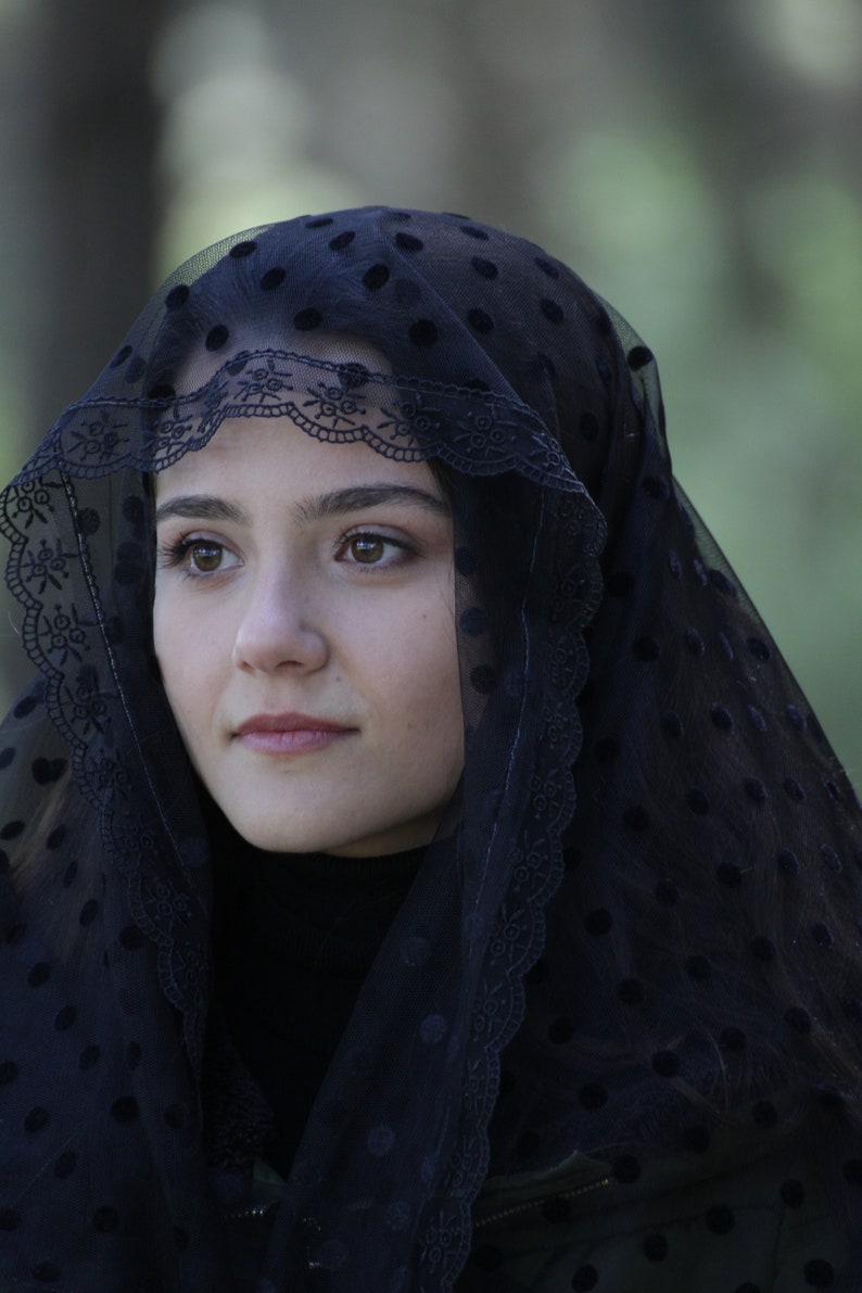 May include: A black veil with a dotted pattern and a decorative lace trim. The veil is draped over the head and shoulders. The person is wearing a black turtleneck.