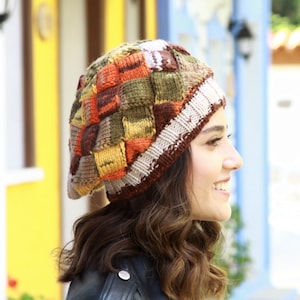 May include: A woman wearing a colorful knitted beret with a patchwork design. The beret is made of brown, orange, yellow, and green yarn.
