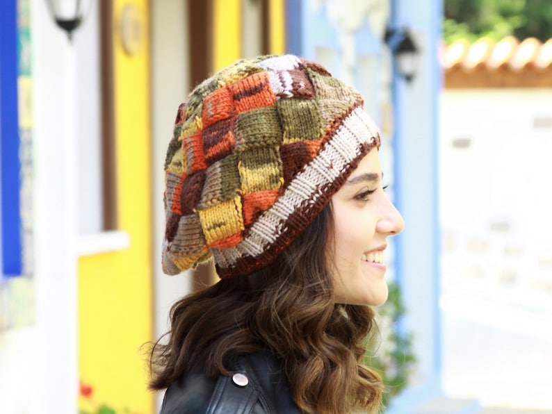 May include: A woman wearing a colorful knitted beret with a patchwork design. The beret is made of brown, orange, yellow, and green yarn.