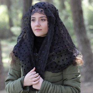 May include: A black veil with a polka dot pattern and lace trim is worn over the head and shoulders. The person is wearing a dark green quilted jacket with a black turtleneck. The background is blurred with trees.