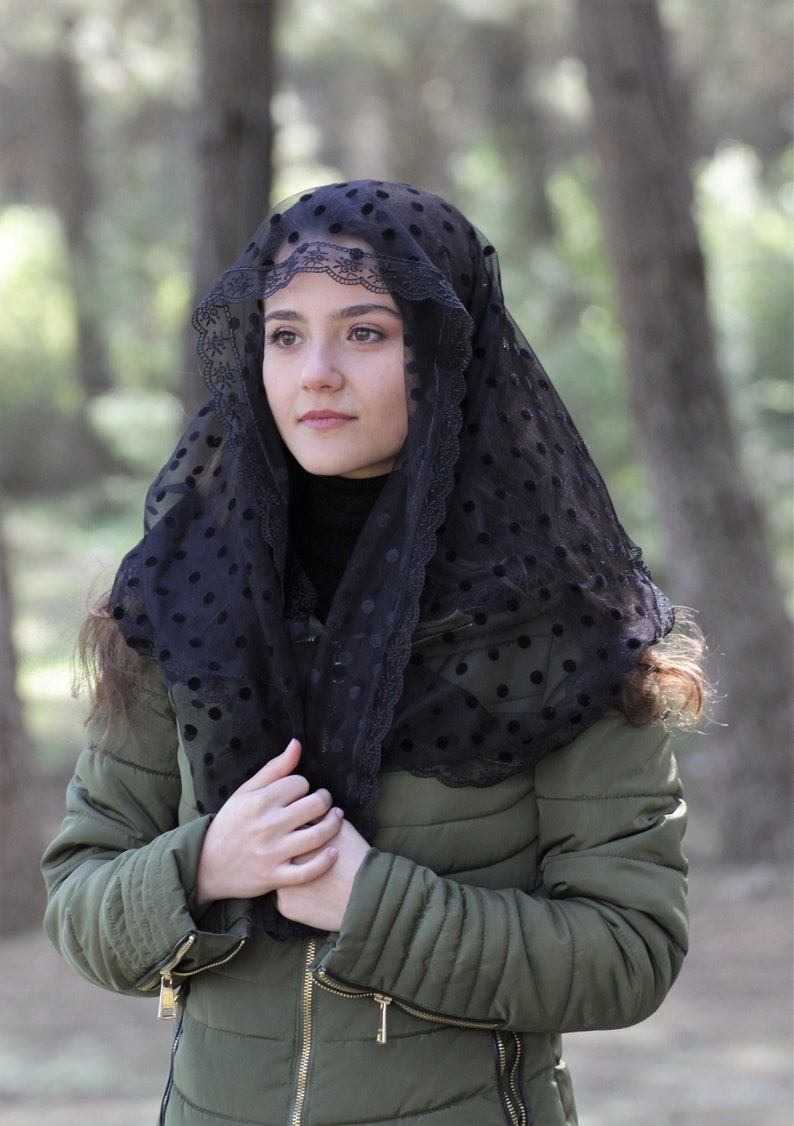 May include: A black veil with a polka dot pattern and lace trim is worn over the head and shoulders. The person is wearing a dark green quilted jacket with a black turtleneck. The background is blurred with trees.
