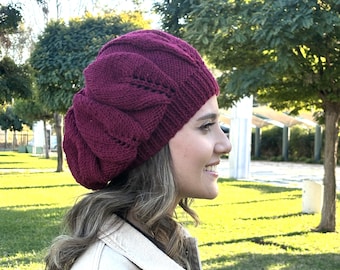 May include: A burgundy knitted beret with a cable knit pattern. The beret is worn by a person with long brown hair.