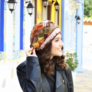 May include: A woman wearing a colorful knitted beanie with a brown leather jacket. The beanie has a patchwork design with brown, orange, yellow, and white yarn.