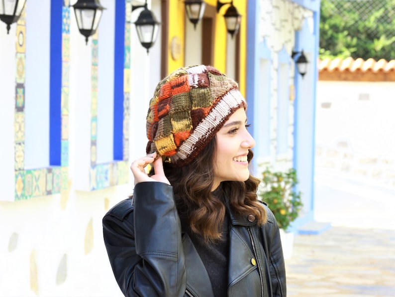 May include: A woman wearing a colorful knitted beanie with a brown leather jacket. The beanie has a patchwork design with brown, orange, yellow, and white yarn.
