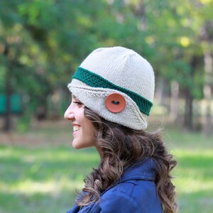 May include: A woman wearing a beige knitted hat with a green band and a brown button. The hat has a unique shape and style.