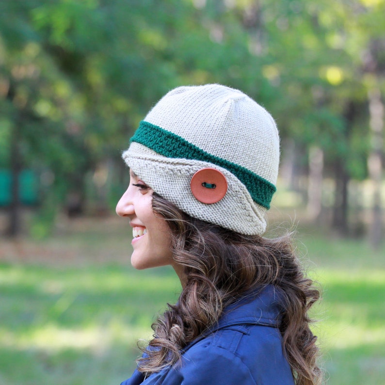 May include: A woman wearing a beige knitted hat with a green band and a brown button. The hat has a unique shape and style.