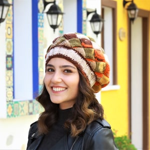 May include: A woman wearing a brown, white, orange, and green patterned knit beanie hat. She is smiling and looking to the right. The background is a colorful wall with windows.