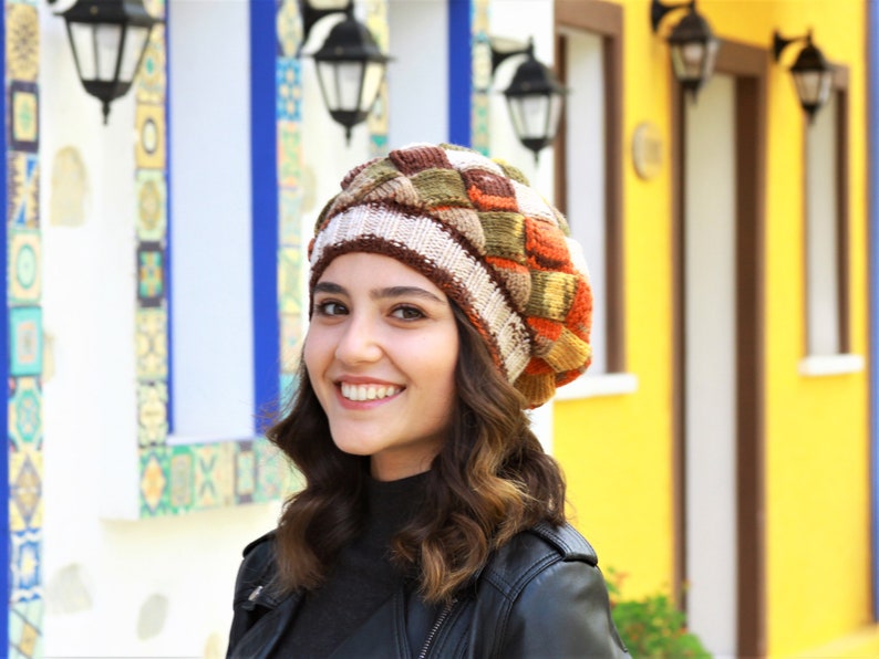 May include: A woman wearing a brown, white, orange, and green patterned knit beanie hat. She is smiling and looking to the right. The background is a colorful wall with windows.