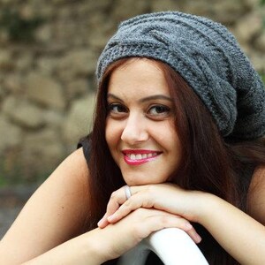 Pode incluir: Um gorro cinza de malha de cabo. O chap&eacute;u &eacute; feito de um material macio e texturizado. A pessoa na imagem est&aacute; sorrindo e usando o chap&eacute;u. O fundo est&aacute; desfocado, com uma parede de pedra e vegeta&ccedil;&atilde;o.