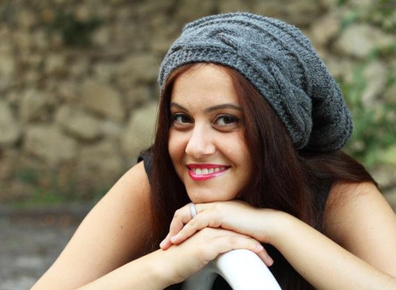 Pode incluir: Um gorro cinza de malha de cabo. O chap&eacute;u &eacute; feito de um material macio e texturizado. A pessoa na imagem est&aacute; sorrindo e usando o chap&eacute;u. O fundo est&aacute; desfocado, com uma parede de pedra e vegeta&ccedil;&atilde;o.