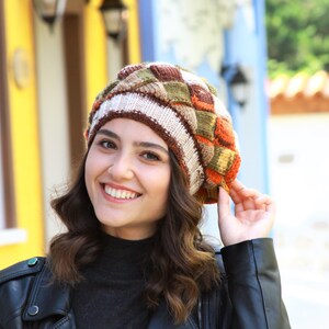 May include: A woman wearing a brown, orange, and green knitted beret with a white stripe. She is smiling and wearing a black leather jacket.