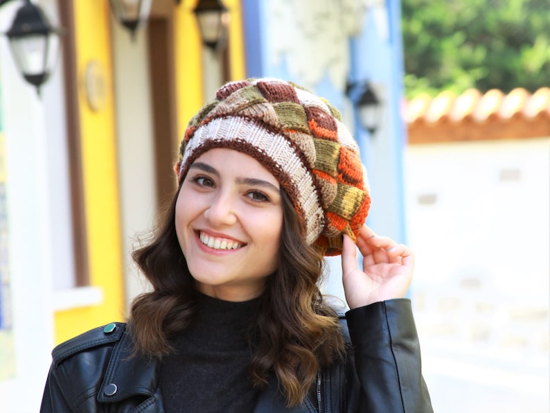 May include: A woman wearing a brown, orange, and green knitted beret with a white stripe. She is smiling and wearing a black leather jacket.