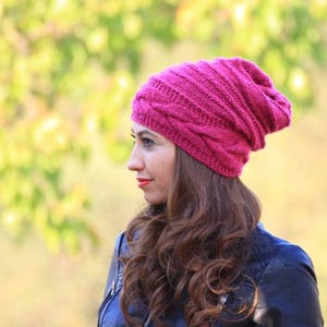 Pode incluir: Um gorro de malha magenta com um design de malha de cabo. O chap&eacute;u &eacute; feito de um material macio e quente, ideal para o clima frio. O chap&eacute;u &eacute; mostrado em uma pessoa com cabelos castanhos longos e ondulados e uma jaqueta preta.