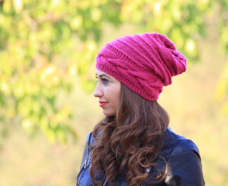 Pode incluir: Um gorro de malha magenta com um design de malha de cabo. O chap&eacute;u &eacute; feito de um material macio e quente, ideal para o clima frio. O chap&eacute;u &eacute; mostrado em uma pessoa com cabelos castanhos longos e ondulados e uma jaqueta preta.
