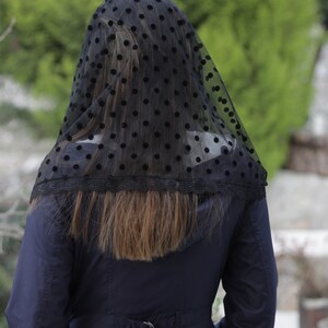 May include: A black veil with a polka dot pattern and a lace trim. The veil is draped over the head and shoulders of a person wearing a navy blue coat. The background is a blurred view of green foliage.