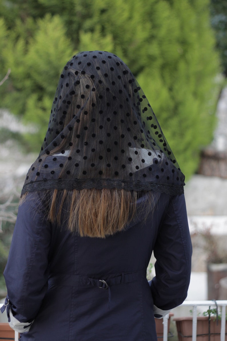 May include: A black veil with a polka dot pattern and a lace trim. The veil is draped over the head and shoulders of a person wearing a navy blue coat. The background is a blurred view of green foliage.
