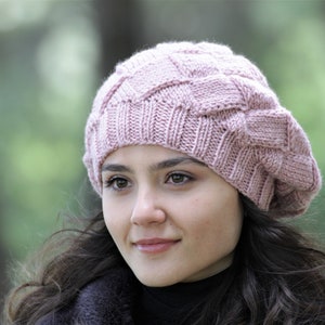 May include: A light pink knitted beret with a textured, basketweave design. The hat is made of soft yarn and has a classic, round shape. This stylish beret is a fashionable accessory for cooler weather.