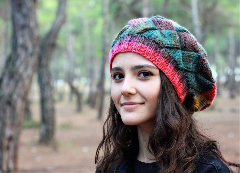 May include: A young person with long brown hair wearing a colourful knitted hat with a pink, green and brown pattern. The hat is a beret style with a wide brim.
