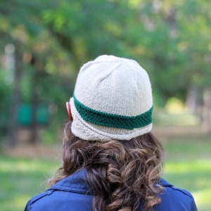 May include: A beige knitted hat with a green band around the crown. The hat has a unique shape with a curved brim.