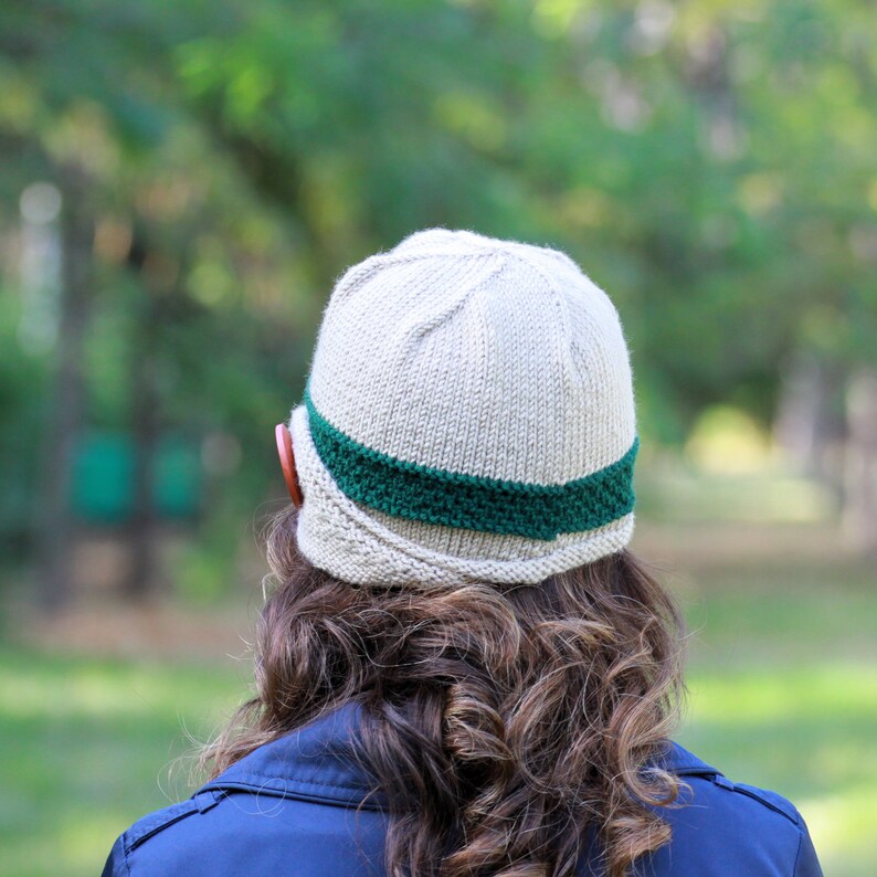 May include: A beige knitted hat with a green band around the crown. The hat has a unique shape with a curved brim.