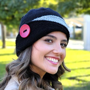 May include: A black and gray knitted hat with a red button. The hat has a wide brim and is angled slightly to the side.
