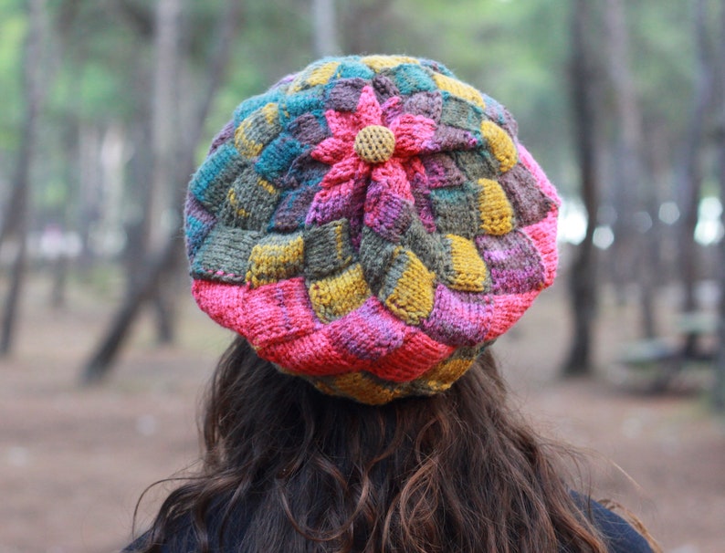 May include: A colourful knitted hat with a flower design. The hat is made of different shades of pink, yellow, green, and brown yarn.