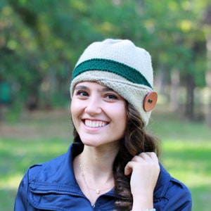 May include: A woman wearing a beige and green knit hat with a brown button. The hat has a wide brim and a green band around the crown. She is smiling and looking at the camera.