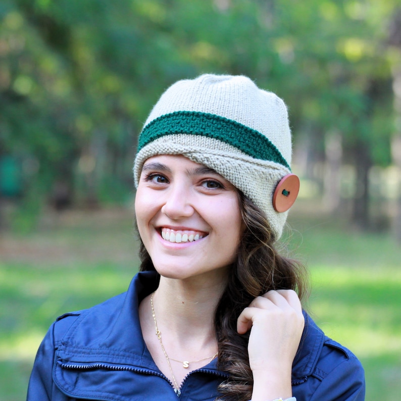 May include: A woman wearing a beige and green knit hat with a brown button. The hat has a wide brim and a green band around the crown. She is smiling and looking at the camera.