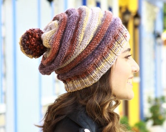 May include: A multi-colored knit beanie with a large pom-pom. The hat features stripes of purple, orange, yellow, and brown. The pom-pom is a dark brown color. The beanie is worn by a person wearing a black leather jacket.