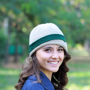 May include: A woman wearing a beige knitted hat with a green band. The hat has a slightly slouched look and covers her forehead. She is smiling and looking at the camera.