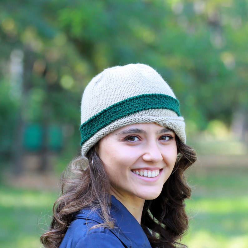 May include: A woman wearing a beige knitted hat with a green band. The hat has a slightly slouched look and covers her forehead. She is smiling and looking at the camera.