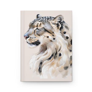 May include: A hardback journal with a watercolour illustration of a snow leopard. The leopard is shown in profile with closed eyes, in shades of white, beige, and black. The journal has a light beige cover.