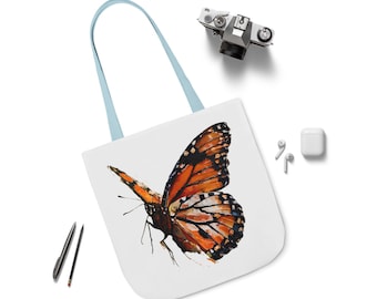Puede incluir: Bolso tote blanco con un llamativo diseño de mariposa naranja y negra. El bolso tiene asas de color azul claro. También se ven una cámara, bolígrafos y auriculares inalámbricos. El bolso está hecho de un material duradero.