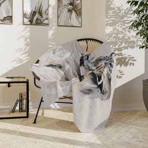 May include: A soft, light-colored throw blanket draped over a black chair. The blanket features a watercolor design with shades of gray, white, and brown, depicting a leopard. The blanket is made of a plush material, perfect for adding warmth and style to any room.