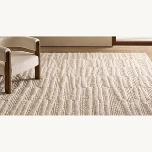 Puede incluir: Alfombra rectangular color crema con un patr&oacute;n de rayas verticales texturizadas. Un sill&oacute;n de color claro con patas de madera se encuentra a la izquierda de la alfombra. La alfombra parece estar hecha de un material suave y afelpado.