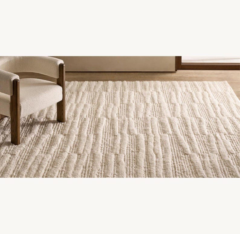Puede incluir: Alfombra rectangular color crema con un patr&oacute;n de rayas verticales texturizadas. Un sill&oacute;n de color claro con patas de madera se encuentra a la izquierda de la alfombra. La alfombra parece estar hecha de un material suave y afelpado.