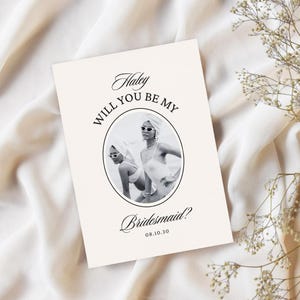 May include: A cream-coloured invitation card with the text "Haley, Will You Be My Bridesmaid?" in elegant script. The card features a black and white photo of two women in sunglasses and headscarves. The date "08.10.30" is also included.