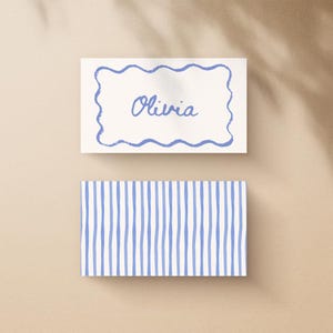May include: Two rectangular cards. The top card has a blue wavy border and the name "Olivia" in blue script. The bottom card has a pattern of vertical blue stripes on a white background. The cards are on a beige surface.
