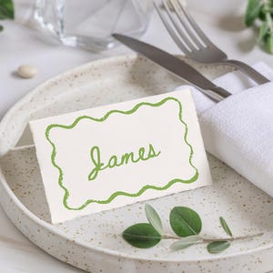 May include: A place setting with a white plate featuring a speckled design. A folded white napkin holds a knife and fork. A place card with a green wavy border and the name "James" is on the plate, along with a sprig of greenery.
