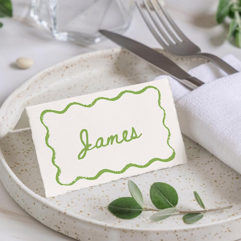 May include: A place setting with a white plate featuring a speckled design. A folded white napkin holds a knife and fork. A place card with a green wavy border and the name "James" is on the plate, along with a sprig of greenery.