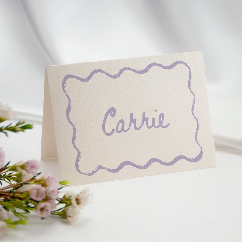 May include: A folded place card with the name "Carrie" in lavender script. The card has a lavender wavy border and is set against a white background. The card is part of a wedding or event stationery set.