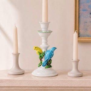 May include: Three white ceramic candle holders with tapered cream-colored candles. The central holder features a sculpted yellow bird and a blue bird perched on green branches. The two side holders have a simple, smooth, flared base design.
