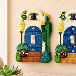 Pode incluir: Duas placas de interruptor de luz com um design moldado de uma porta arqueada azul com um interruptor de alavanca branco no centro. As placas são decoradas com cactos verdes, flores amarelas, um sino preto e um pequeno vaso castanho.