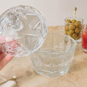 May include: A clear glass bowl with a matching lid featuring a faceted starburst pattern. The bowl has an octagonal shape with thick walls. Nearby are three small glass containers filled with green olives, red cherries, and yellow lemon slices.