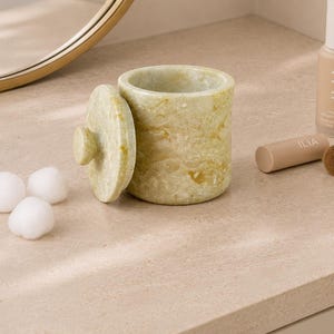 May include: A cylindrical light green stone container with a matching circular lid featuring a small knob handle. The container is accompanied by three white cotton balls, a bottle of Ilia foundation, a lipstick tube, and a makeup brush.