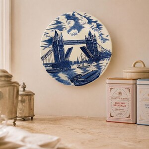 May include: A round ceramic decorative plate featuring a blue and white illustration of Tower Bridge in London with a boat on the water. The artwork includes detailed architectural lines of the bridge towers and suspension cables against a cloudy sky.