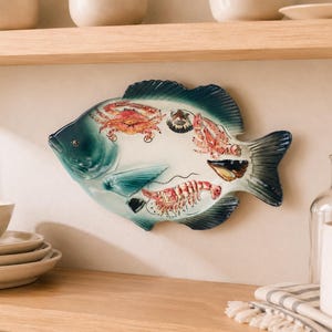 May include: A fish-shaped ceramic serving platter featuring a gradient teal and blue finish. The center of the platter is decorated with detailed illustrations of a red crab, a shrimp, and various seashells in shades of orange, brown, and white.