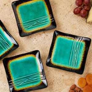 May include: Four square glass plates featuring a vibrant turquoise centre that fades into a dark black border with a golden-brown transition. Each plate is decorated with a series of thin, white, parallel lines that cross over one another.