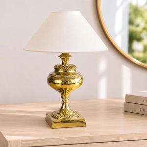 May include: A table lamp with a polished gold-tone metal base featuring a tiered, rounded body and a square pedestal. The lamp is topped with a plain, off-white conical fabric shade that provides a neutral light diffusion for the fixture.