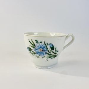May include: White teacup with a blue floral design. The cup features a gold rim and a handle. The floral pattern includes a large blue flower with green leaves and buds. A classic design for tea or coffee.